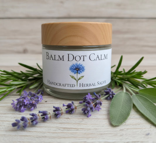 A 2-ounce glass jar of Balm Dot Calm herbal salve with a light wood lid and white label, styled on a light wood surface with lavender sprigs, rosemary, and sage leaves.