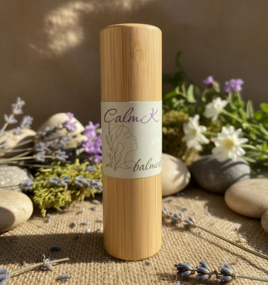 Balm Dot Calm's Calm Kiss Balm in a natural bamboo tube, nourishing botanical lip balm photographed on a soft neutral background upright on a burlap surface surrounded by lavender sprigs, small flowers, and smooth stones in warm natural lighting.