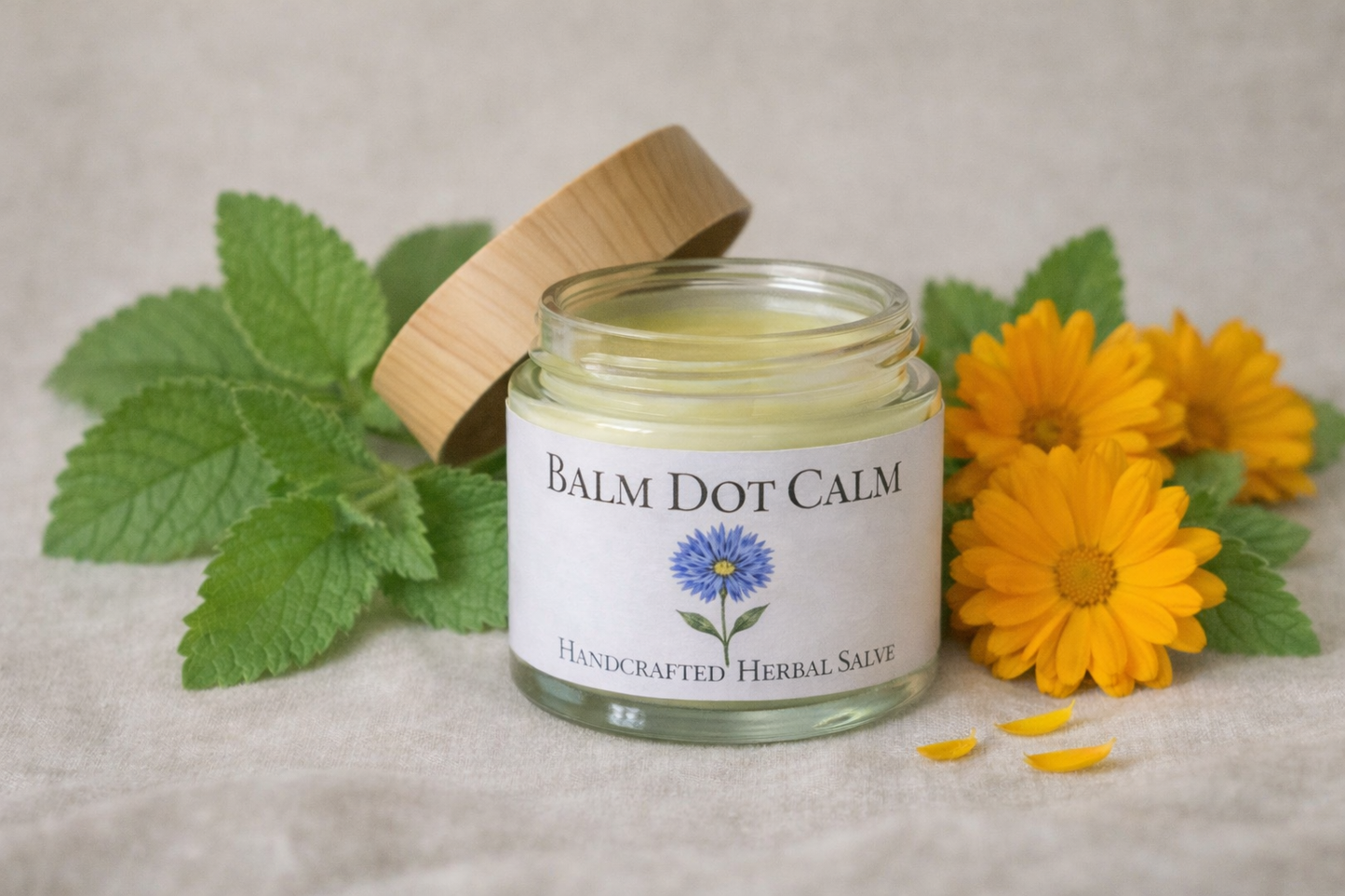 A 2 oz glass jar of Balm Dot Calm Essential Calm herbal salve with a bamboo lid, styled on neutral fabric with green leaves and yellow calendula petals.